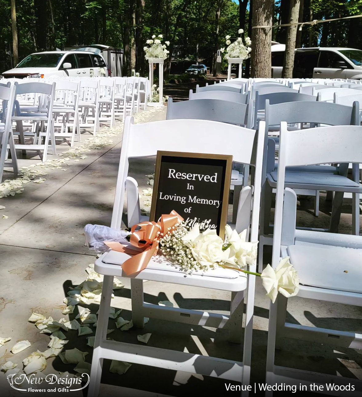 An outdoor aisleway of White padded folding chairs, on pavement. the centere aisle lined with white rose petals. A memorial chalkboard sits atop the foremost chair, with a bouquet of white roses placed lovingly at the front.